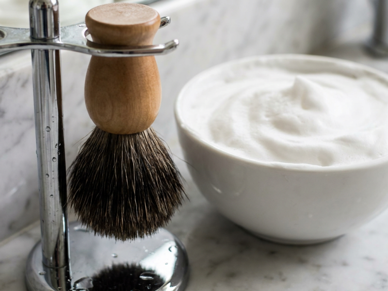 How to clean a shaving brush: wet badger brush hanging bristles down on a chrome stand beside a lather bowl