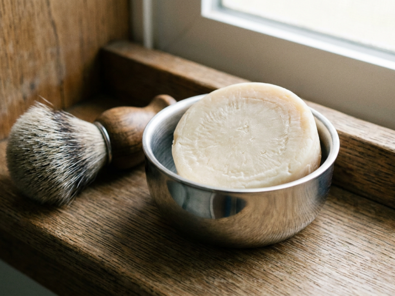 Round shaving soap puck seated in an open stainless steel shaving bowl on a wooden shelf