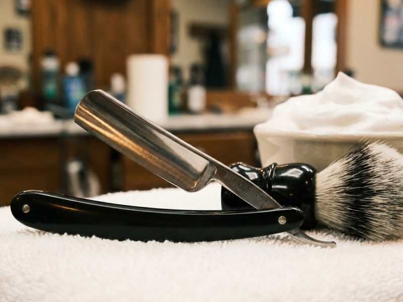 Straight razor head shave with an open straight razor beside a shaving brush and bowl of lather on a barbershop counter