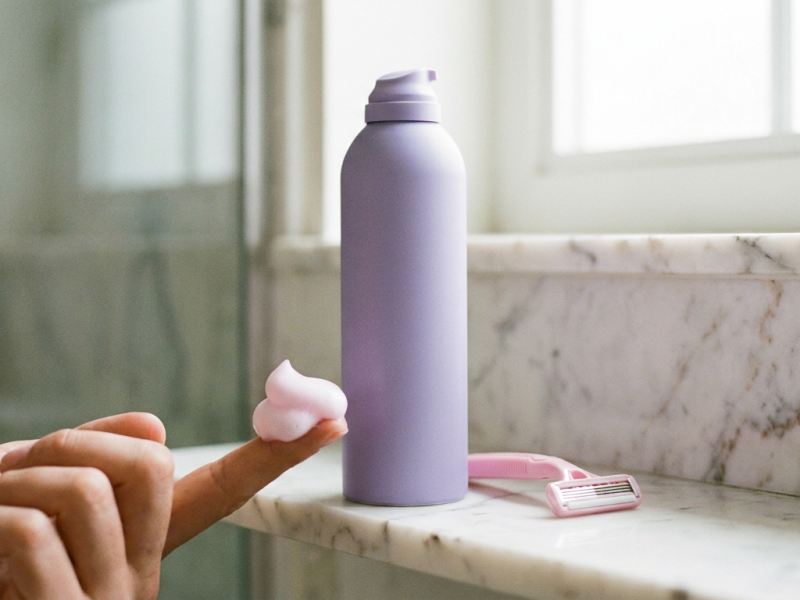 Skintimate shave gel lavender aerosol can with foam and a pink razor on a bathroom shelf